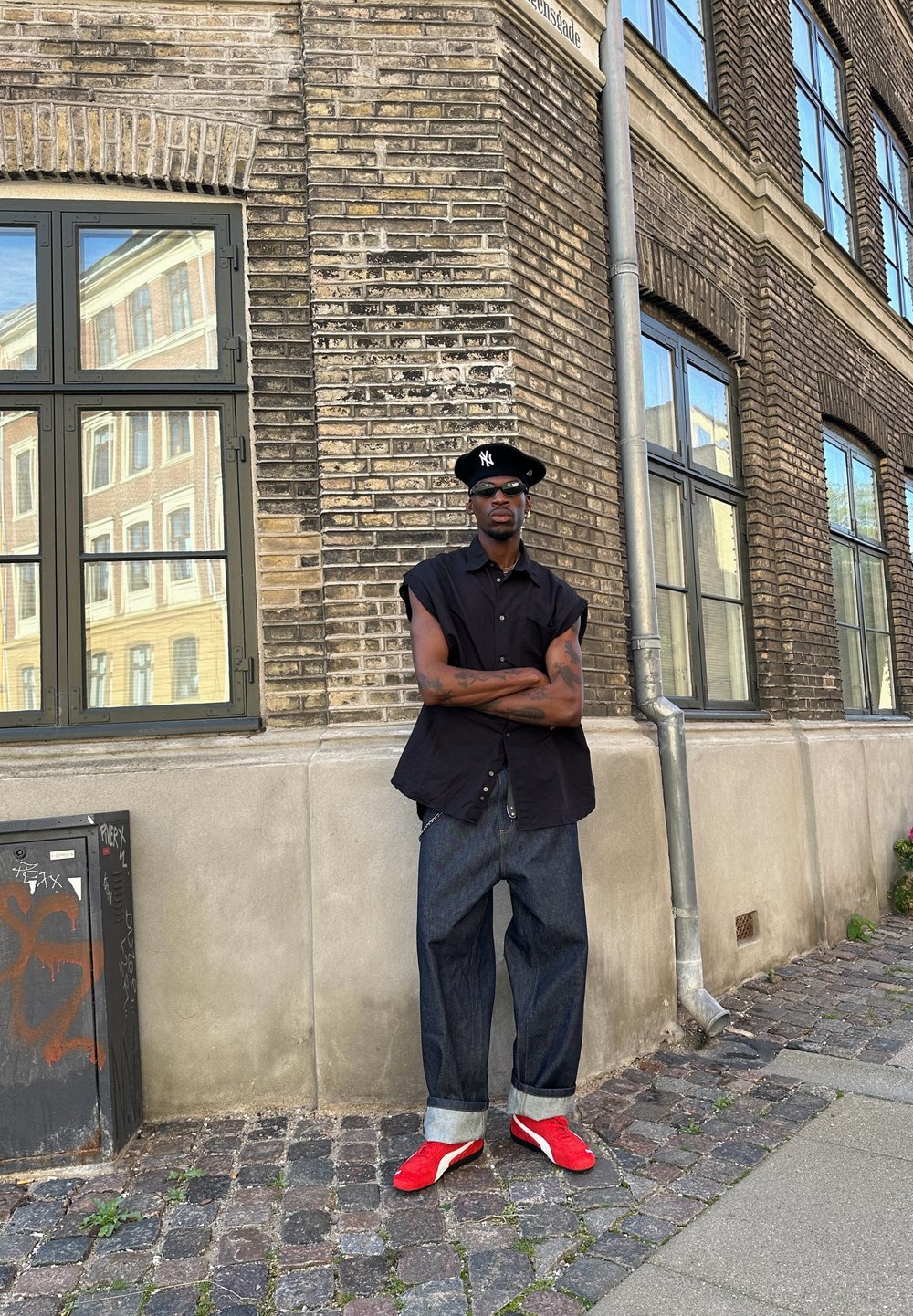 Μαύρο αμάνικο μπλουζάκι, ξεθωριασμένα τζιν παντελόνια, κόκκινα αθλητικά παπούτσια. Στηρίζεται σε τοίχο από τούβλα με παράθυρα, με τα χέρια σταυρωμένα, γυαλιά ηλίου και καπέλο.