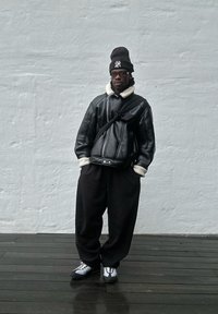 Giacca in pelle nera con colletto in shearling bianco, pantaloni neri larghi e berretto nero. Sneakers bianche e grigie con lacci. Sfondo in legno.