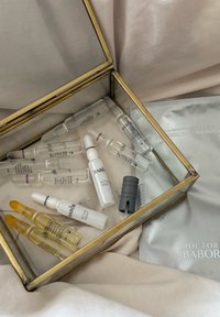 Hudpleieampuller merket BABOR, inkludert Active Night, inni en glassboks med et grått lokk og Doctor BABOR øyeområde lapper på beige stoff.