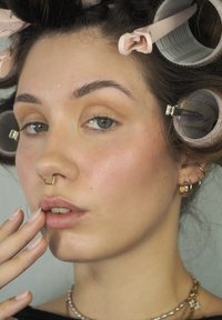 Gros plan sur un visage avec des yeux verts, une peau claire et des bijoux de septum en or. Cheveux en rouleaux roses, lèvres brillantes, ongles manucurés dans une couleur pâle.