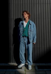 Jeansjacke über einem grünen Cardigan mit Holzknöpfen, weißem T-Shirt, hellblauen Jeans und grauen Sneakers mit einem weißen Swoosh-Detail.