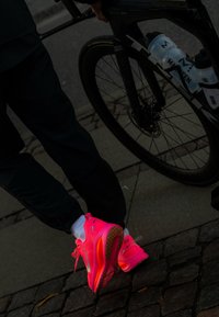 Personne portant des chaussures de course rose vif et des chaussettes blanches debout à côté d'un vélo de course noir sur une surface pavée.
