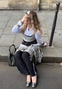 Fiatal nő hosszú, hullámos hajjal, városi járdaszegélyen ülve, réteges ruházatot, fekete nadrágot, magassarkút visel, egyik kezével a fejét tartja.