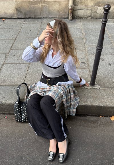 Joven con el cabello largo y ondulado sentada en la acera de la ciudad, vestida con ropa superpuesta, pantalones negros, tacones y sosteniendo su cabeza con una mano.