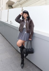 Giacca cropped in patent leather marrone sopra una mini gonna a pieghe grigia, abbinata a stivali neri a punta alti fino al ginocchio e a una borsa nera.