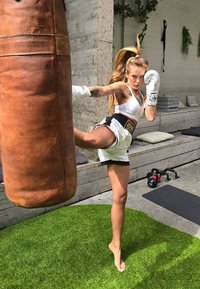 Πυγμάχος με λευκά γάντια και μπλουζάκι crop top ρίχνει μια κλωτσιά σε ένα καφέ δερμάτινο σάκο πυγμαχίας σε τεχνητό γρασίδι. Κονταροκρέπτες είναι ορατοί στο φόντο.