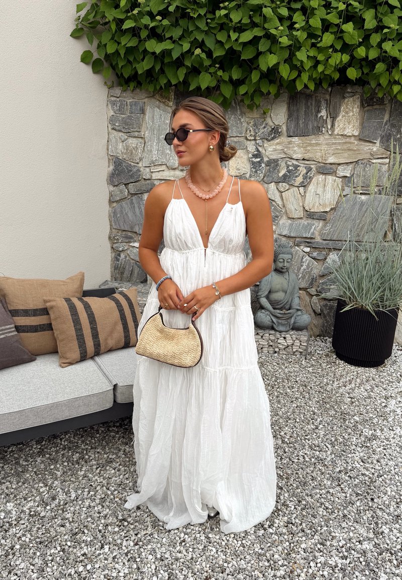 Femme en robe blanche fluide tenant un sac tressé, debout sur du gravier près d'un mur en pierre et un canapé d'extérieur avec des coussins marron.