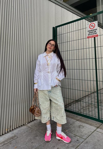 Camisa blanca con cuello y lazos laterales, pantalones cortos de mezclilla con agujeros, zapatillas rosas y un bolso estampado. El look se completa con pelo largo y gafas.