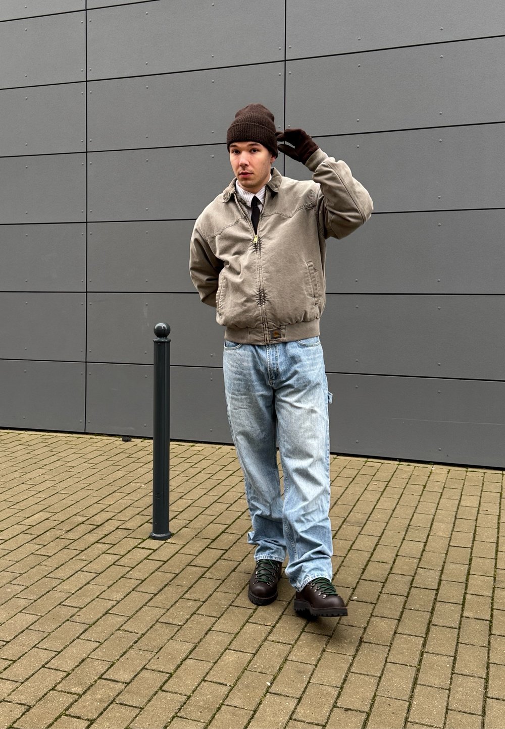 Καφέ μπουφάν bomber πάνω από λευκό πουκάμισο και μαύρη γραβάτα, μπλε τζιν και καφέ μπότες με πράσινα κορδόνια; στέκεται κοντά σε μία μαύρη κολώνα, με γκρίζο τοίχο πίσω.