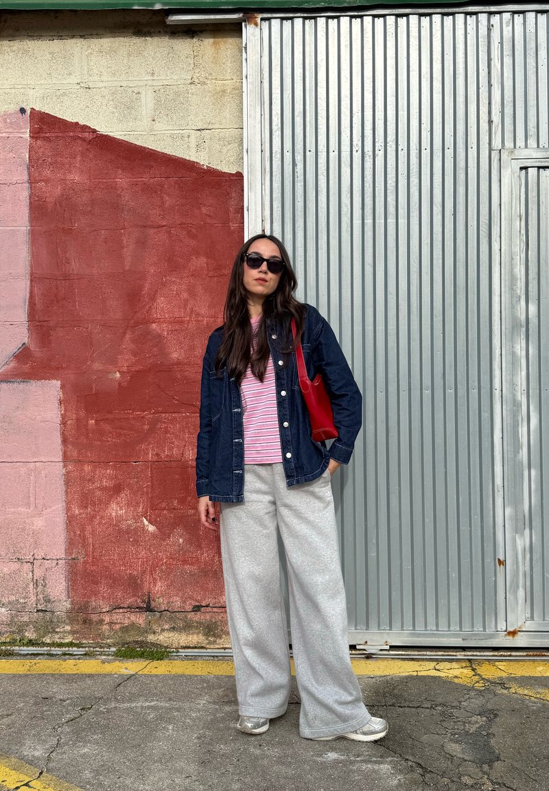 Spijkerjasje, gestreept roze-wit T-shirt, grijze joggingbroek en rode tas. Zwarte zonnebril; achtergrond met rode en grijze muren.