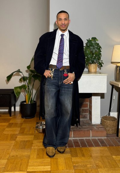 Hombre con un abrigo largo negro, camisa blanca, corbata azul con estampado, vaqueros de pierna ancha con llaveros y zapatos color marrón claro, de pie en una habitación con plantas y una chimenea.