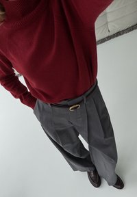 Weinroter übergroßer Pullover, graue plissierte Hose mit braunem Gürtel und goldener ovaler Schließe, sowie dunkelbraune Loafers. Glatte Texturen, moderner Schnitt.