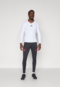 Balta garām piedurknēm sporta krekls ar melnu logotipu, kombinēts ar pielāgotām pelēkām biksēm ar sarkanu logotipu un svītrām, valkātas ar melnām kurpēm.