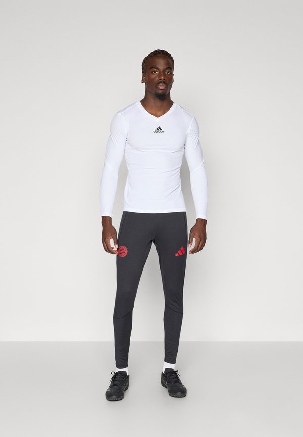 Balta garām piedurknēm sporta krekls ar melnu logotipu, kombinēts ar pielāgotām pelēkām biksēm ar sarkanu logotipu un svītrām, valkātas ar melnām kurpēm.