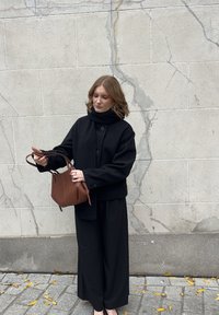 Vrouw in een zwarte jas en wijde broek houdt een bruine handtas vast terwijl ze op het trottoir staat, omringd door verspreide gele bladeren bij een gebarsten stenen muur.