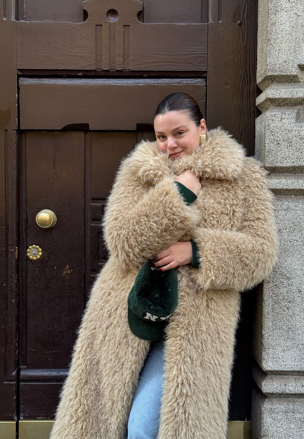 Cappotto lungo e soffice color beige con un ampio colletto e una texture morbida, indossato sopra un maglione verde. Una berretto verde è tenuto in una mano.