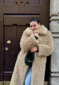 Cappotto lungo e soffice color beige con un ampio colletto e una texture morbida, indossato sopra un maglione verde. Una berretto verde è tenuto in una mano.