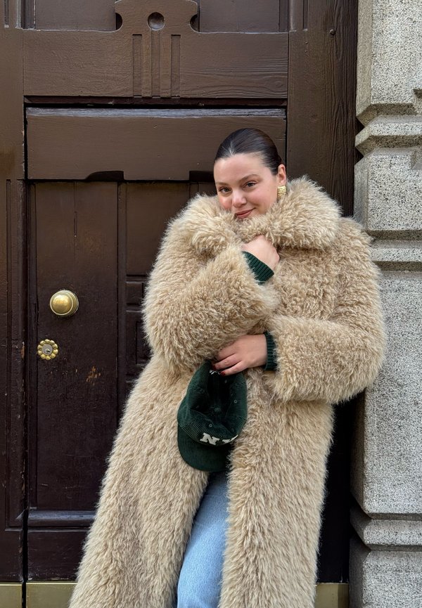 Beżowy, puszysty długi płaszcz z szerokim kołnierzem i miękką fakturą, noszony na zielonym swetrze. Zielona czapka trzymana jest w jednej ręce.