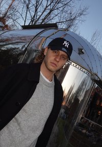 Una persona che indossa un berretto nero, un maglione grigio e un cappotto nero si trova accanto a una superficie argentata riflettente con alberi sullo sfondo.