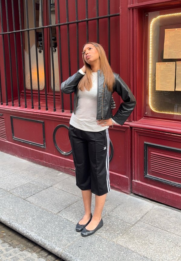 Jeune femme en veste en cuir noire et pantalon de survêtement court se tient sur le trottoir près d’un bâtiment rouge avec des fenêtres grillagées et un affichage de menu.