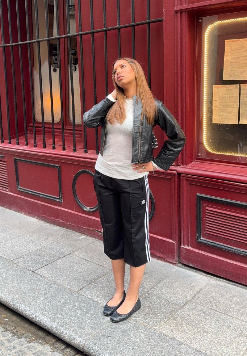 Jeune femme en veste en cuir noire et pantalon de survêtement court se tient sur le trottoir près d’un bâtiment rouge avec des fenêtres grillagées et un affichage de menu.