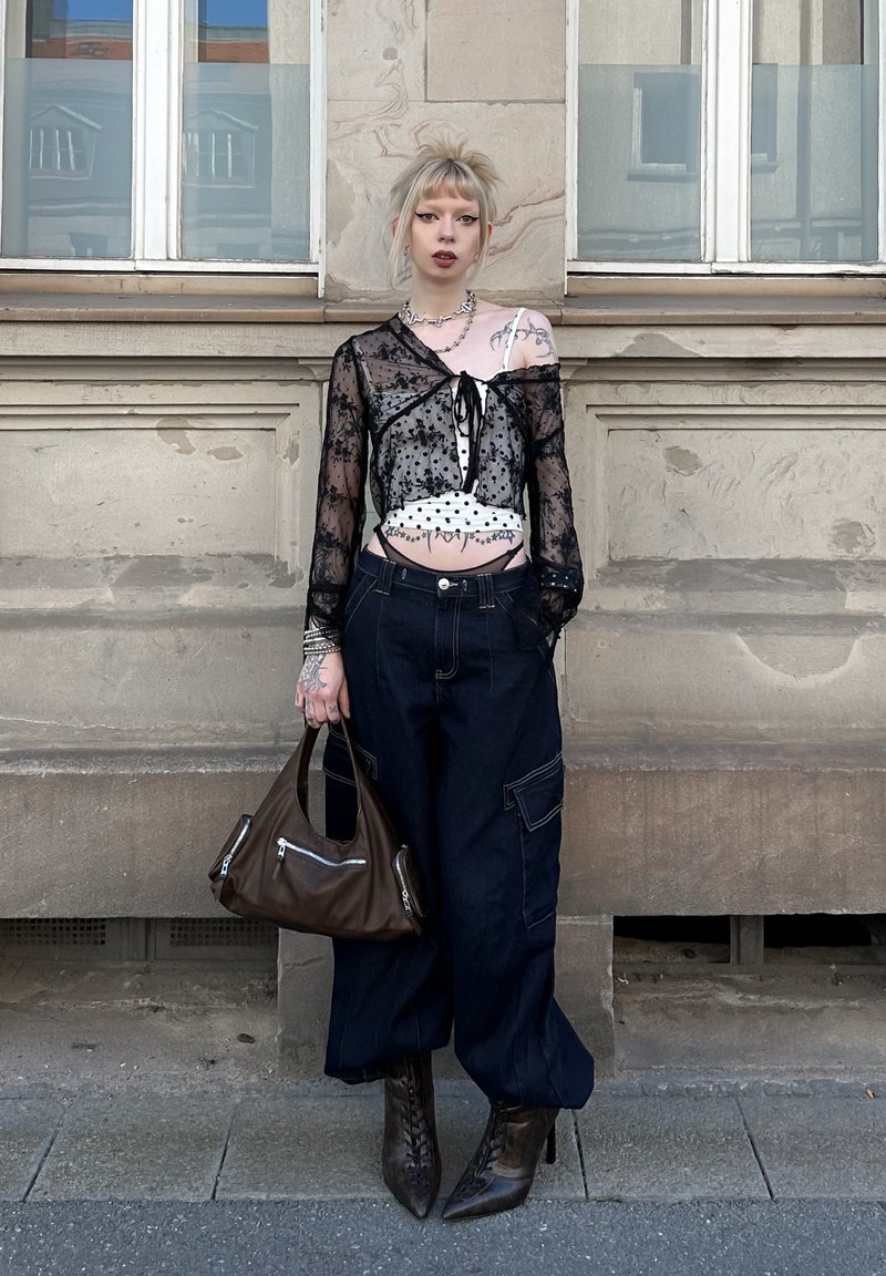 Personne aux cheveux blonds portant un haut transparent noir par-dessus un crop top blanc à pois, pantalon ample foncé et bottes à talons hauts, tenant un sac à main marron dans la rue.