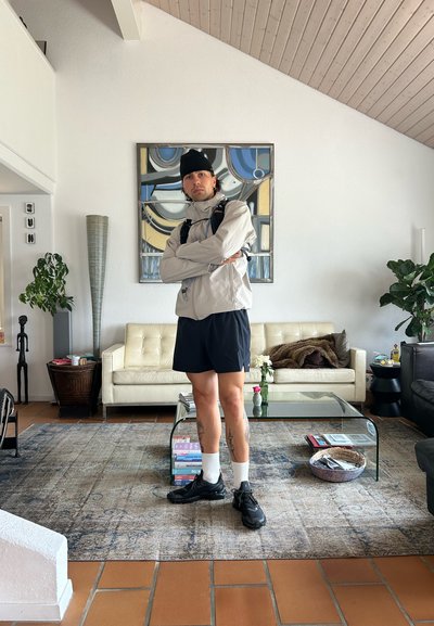 Chaqueta impermeable gris claro, pantalones cortos negros y zapatillas negras usados en una sala de estar moderna con una mesa de café de cristal y plantas decorativas.