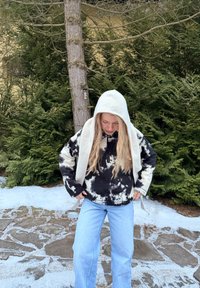 Femme aux longs cheveux blonds portant une veste à motifs noir et blanc, un jean bleu clair et une capuche blanche, debout sur un pavé en pierre légèrement enneigé en plein air.