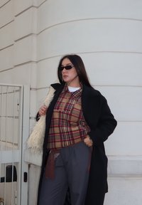 Femme portant un manteau noir sur une chemise à carreaux rouge et un pantalon gris, tenant un sac blanc duveteux, debout contre un mur blanc incurvé.
