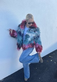 Pūkains daudzkrāsains mākslīgās kažokādas Žakete ar sarkaniem, ziliem un rozā toņiem, apvienota ar gaiši ziliem platsvārkiem džinsiem un brūnām apaviem ar kniedēm.