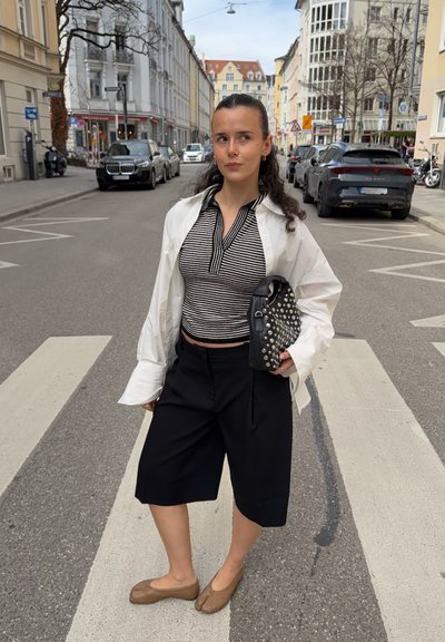 Joven mujer está en un paso de peatones de la ciudad, lleva un top de rayas, una camisa blanca abierta, pantalones cortos negros y zapatos marrones claros, sosteniendo un bolso negro con tachuelas.