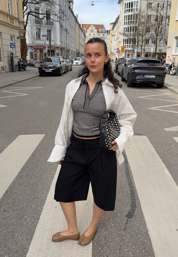 Mladá žena stojí na mestskom priechode pre chodcov, má na sebe pruhovaný top, bielu otvorenú košeľu, čierne šortky a béžové topánky, drží čiernu kabelku s cvokmi.