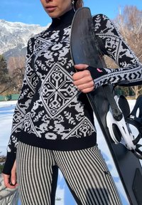 Femme portant un pull à motifs noirs et blancs et un pantalon rayé tenant un snowboard en plein air, avec de la neige et des montagnes en arrière-plan.
