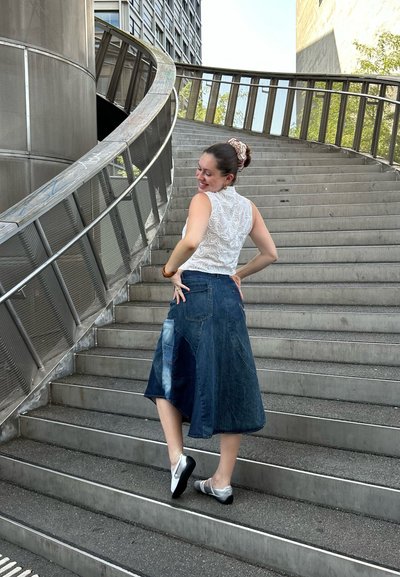 Falda de mezclilla con dobladillo asimétrico, parches en azul claro y oscuro, combinada con una blusa de encaje blanca. La modelo lleva zapatos grises y negros, en las escaleras.