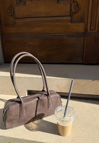 Brun semsket veske med to håndtak og glidelås, plassert ved siden av et klart glass med iskaffe på en lys steinflate.