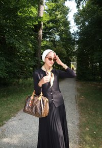 Barna Louis Vuitton kézitáska arany kiegészítőkkel, fekete gombos kardigán, sárga sál és hosszú fekete pliszírozott szoknya erdős környezetben.