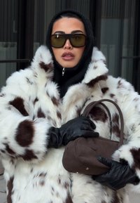 Műszőrméből készült kabát fehér és barna foltmintákkal, fekete kesztyűkkel és egy kis barna szarvasbőr kézitáskával. Magas gallérral és cipzáras lezárással rendelkezik.