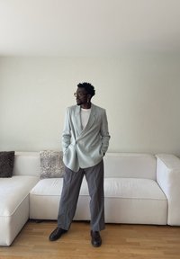 Muž v svetlomodrom saku, bielej košeli, sivých nohaviciach a tmavých topánkach stojaci s rukami v vreckách pred bielou pohovkou s vankúšmi.