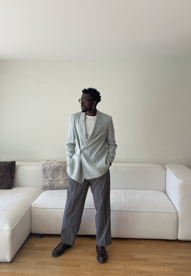 Man wearing light blue blazer, white shirt, gray pants, and dark shoes, standing with hands in pockets in front of white sofa with cushions.