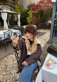 En person med en brun pelskasket og en sort frakke sidder ved et bord med en cappuccino, omgivet af grønt og havemøbler.