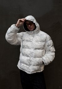 Hvit dunjakke med teksturert, mønstret overflate, høy krage og glidelås foran. Modellen har på seg solbriller og har hendene i lommene.