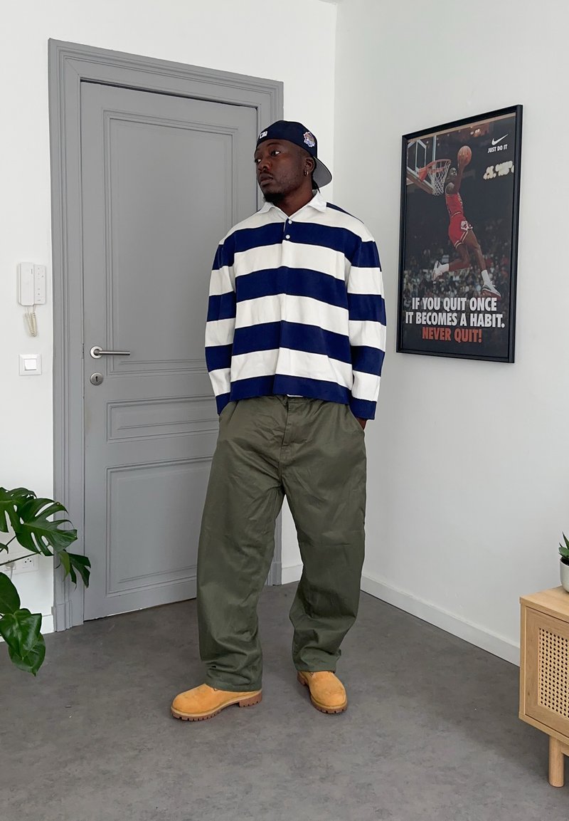 Mann mit blau-weiß gestreiftem Hemd, grüner Hose und hellbraunen Stiefeln steht neben einer grauen Tür und einem motivierenden Basketballposter an der Wand.