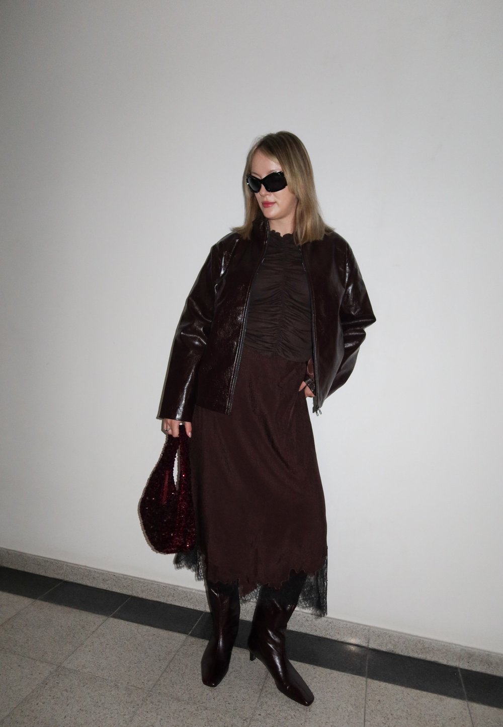 Femme portant une veste en cuir marron foncé, un haut froncé, une longue jupe, des bottes montant au genou, des lunettes de soleil, et tenant un sac à main à sequins rouge foncé contre un mur uni.