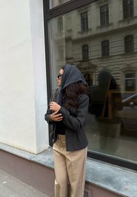 Femme portant un foulard gris, un blazer sombre, un pantalon beige et des lunettes de soleil, debout dans une rue de la ville près d'une fenêtre reflétant un bâtiment historique.