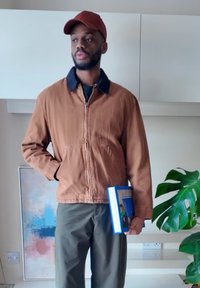 Brown zip-up jacket with a blue collar, olive green trousers, holding a blue book, standing near a plant against a neutral background.