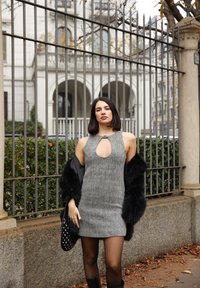 Γκρίζα πλεκτή φόρμα με στρογγυλή κοπή στο στήθος, συνδυασμένη με ένα μαύρο γούνινο παλτό. Κρατάει μια μαύρη τσάντα με καρφιά, μπροστά από ένα πλέγμα φράχτη.