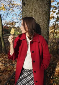 Palton roșu din lână cu nasturi mari negri, pulover tricotat crem și fustă cu carouri. Ținând o frunză galbenă, stând lângă un trunchi de copac cu textură.