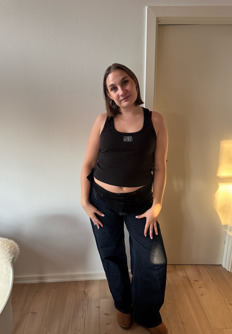 Giovane donna con capelli castani corti, che indossa una canotta nera e jeans scuri a gamba larga, in piedi all'interno contro una parete bianca e una porta chiusa.