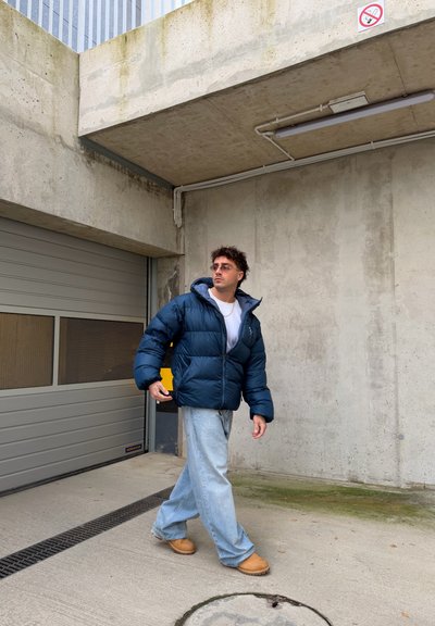 Chaqueta acolchada en azul marino, con capucha y cierre de cremallera. Combinada con vaqueros acampanados de azul claro y calzado marrón claro. Fondo de hormigón.