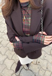 Femme portant un blazer brun foncé et un col roulé à carreaux, collier en or et bagues, tenant un sac à bandoulière à chaîne, debout sur un trottoir à motifs.